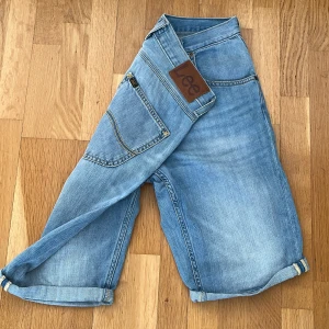 Ljusblå jeansshorts från Lee - Snygga ljusblå jeansshorts från Lee med klassisk femficksdesign och uppvikta benslut. Shortsen har Lee-lapp bak i midjan och gul kontrastsöm. Perfekta för en avslappnad stil.
