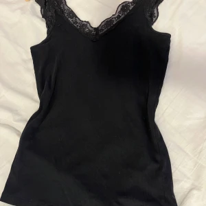 Svart linne med spets - Svart ribbad linnetopp med v-ringning och spetsdetaljer längs halsringningen. Perfekt att styla till olika outfits och superskön! 