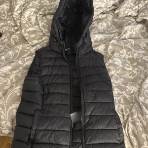 Svart dunväst från ONLY - Säljer en svart dunväst från ONLY i storlek XS. Västen har huva och dragkedja framtill, perfekt att slänga över en tröja för extra värme. Snygg och enkel design som passar till mycket.