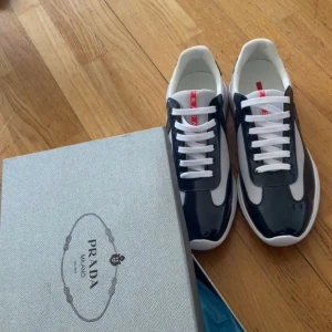 Prada American Cup’s  - Säljer ett par stilrena sneakers från Prada i marinblå och vitt med röda detaljer på plösen och hälen. Skorna har vit sula, snörning och en kombination av glansigt material och mesh. Perfekta för dig som vill ha en lyxig och sportig look. Aldrig använt de, då de är väldigt tighta i storleken, så den passar perfekt om du har 46 i normala skor.