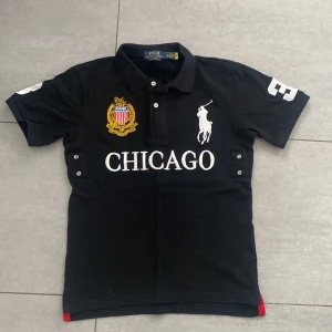 Ralph Lauren Chicago polo tröja - Tröajn är 65 cm lång och 46 cm bredd.Tröjan är en svart pikétröja med ordet "CHICAGO" skrivet i stora vita bokstäver. Den har även siffran "3" i vitt på högerärmen, en logotyp på övre bröstet och en Polo-logotyp på höger sida.