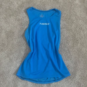 Blå träningstopp från Fusion - Säljer en blå ärmlös träningstopp från Fusion i funktionsmaterial. Toppen har rund halsringning och meshpaneler på sidorna för extra ventilation. Perfekt för löpning eller gympass!