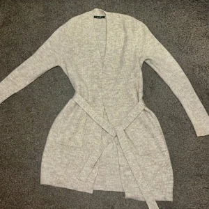 Ljusgrå ribbad kofta med bälte - Mysig ljusgrå kofta med ribbad struktur och knytskärp i midjan. Koftan har långa ärmar och två stora fickor framtill. Perfekt att slänga på sig för en avslappnad och stilren look👚🩶