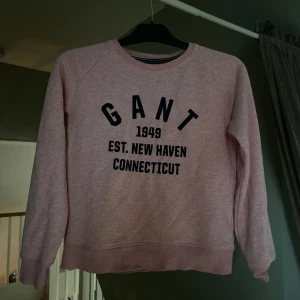 Ljusrosa sweatshirt från GANT - Ljusrosa sweatshirt från GANT med svart tryck framtill: 'GANT 1949 EST. NEW HAVEN CONNECTICUT'. Klassisk rund halsringning och långa ärmar. Mjuk bomullskvalitet. En liten fläck några cm under texten som inte alls märks i verkligen. Såg det enbart på bilden