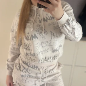 Dress - Säljer en krämvit grå dress graffiti-inspirerat tryck över hela dressen. Tröjan har huva med  och långa ärmar samt ribbade muddar. Perfekt för en avslappnad och trendig look.