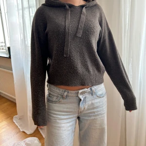 Mörkgrå croppad hoodie - Säljer en mörkgrå croppad hoodie med långa ärmar och dragsko i huvan. Tröjan är stickad och har en avslappnad passform, perfekt till jeans eller mjukisbyxor. Superenkel att styla för en chill look.