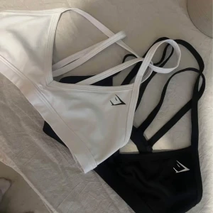 Sportiga linne-toppar från Gymshark - Två stilrena linne-toppar från Gymshark, en i vitt och en i svart. Båda har smala axelband och en minimalistisk logga framtill. Perfekta för träning eller en sportig look. 150kr styck