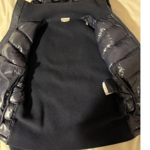 Svart dunväst med stickade ärmar från Moncler - Snygg svart dunväst från Moncler med glansig quiltad framsida och huva. Västen har stickade ärmar och ribbad rygg, samt dragkedja framtill. Moncler-logga på ärmen och praktisk ficka. Perfekt för lager-på-lager-stil.