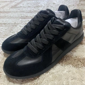 Svarta sneakers från Maison Margiela - Säljer ett par svarta sneakers från Maison Margiela med en mix av mocka och skinn. Skorna har rund tå, platt sula och klassisk snörning. Perfekta för dig som gillar stilrena och tidlösa sneakers med exklusiv känsla.