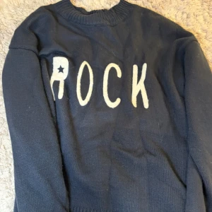 Mörkblå stickad tröja med texten ROCK - Säljer en mörkblå stickad tröja med vit text 'ROCK' framtill. Tröjan har rund halsringning och långa ärmar. Perfekt för dig som gillar en cool och avslappnad stil.