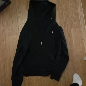 Marinblå hoodie från Ralph Lauren - Marinblå hoodie från Ralph Lauren med dragkedja framtill och klassisk broderad logga på bröstet. Tröjan har långa ärmar och huva, perfekt för en avslappnad stil. Passar både killar och tjejer som gillar stilrena plagg.