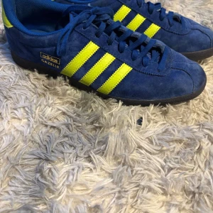 Adidas Gazelle blå och gula sneakers - Säljer ett par Adidas Gazelle sneakers i blå mocka med gula ränder och detaljer. Skorna har klassisk snörning och svart platt sula. Perfekta för dig som gillar retrostil och vill sticka ut med färg.