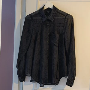 Svart transparent skorta/blus från R&D! - Säljer en svart transparent skjorta från R&D med diskret ormmönster. Fina veck i ryggen som för att det blir fint fall på den. (Liten oklar fläck som kanske/kanske inte går bort i maskinen. Se sista bilden)