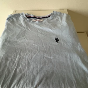 Ljusblå t-shirt från U.S. Polo sport - Säljer en ljusblå t-shirt från U.S. Polo sport. med klassisk rund hals och broderad logga på bröstet. T-shirten har korta ärmar och är tillverkad i mjuk bomull. Perfekt för en avslappnad stil. Har ett litet hål på ryggen. Men inga andra skador.
