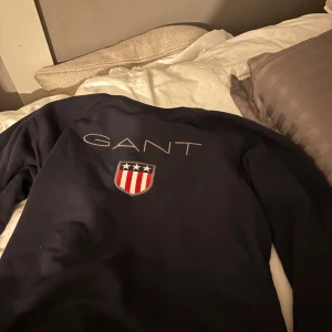 Mörkblå sweatshirt från GANT - Säljer en mörkblå sweatshirt från GANT med stort broderat GANT-tryck och sköld med stjärnor och ränder på ryggen. Perfekt för en stilren och sportig look. Klassisk passform och långärmad modell.