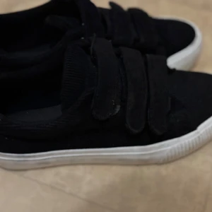 Svarta sneakers med kardborreband - Säljer ett par svarta sneakers med vit sula och tre breda kardborreband. Skorna har en klassisk rund tå och är perfekta för en stilren vardagslook. Materialet är syntet och de är lätta att ta på och av. Väldigt fint till vardag och annat.