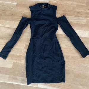 Svart kortklänning med snörning och cut-out axlar - Säljer en svart kortklänning med lång ärm och coola cut-out detaljer vid axlarna. Klänningen har hög hals och snygg snörning i ryggen som ger en edgy look. Perfekt för dig som gillar unika detaljer och vill sticka ut.
