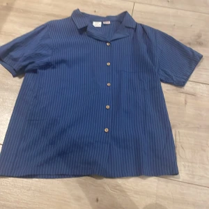 Blå randig skjorta från Zara - Säljer en blå kortärmad skjorta från Zara med diskreta vertikala ränder och knappar framtill. Skjortan har klassisk krage och bröstficka, perfekt för en avslappnad stil.