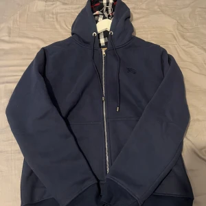 Burberry hoodie  - Säljer en marinblå hoodie från Burberry med dragkedja och klassiskt rutigt foder i huvan. Tröjan har broderad logga på bröstet och justerbar huva med snören. Perfekt för en stilren och bekväm look.
