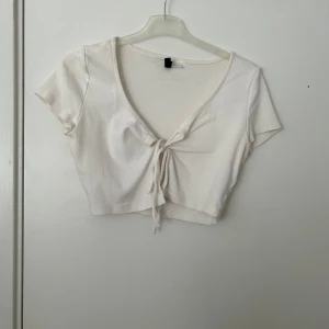 Vit croppad knyt-topp - Säljer en vit croppad topp med korta ärmar och v-ringning. Toppen har en snygg knytning framtill som ger en trendig look. Perfekt att matcha med höga jeans eller kjol.