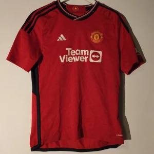 Manchester United matchtröja - Röd Manchester United fotbollströja från Adidas med klubbmärke på bröstet och TeamViewer sponsortryck. Tröjan har svarta detaljer på axlar och sidor samt vita Adidas-logga. Klassisk rund hals och korta ärmar. Perfekt för fans! Storlek S. Obs inget namn eller nummer på ryggen. Kolla profilen för mer fotbollströjor och grisch kläder bland annat .