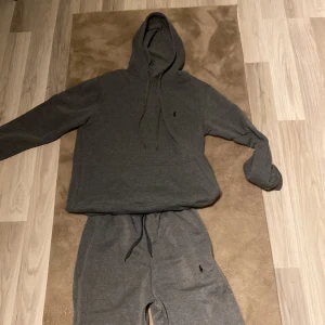 Grå tracksuit från raulph lauren - Mysig grå tracksuit från Ralph Lauren med klassisk huva och dragsko. Tröjan har långa ärmar och en liten broderad logga på bröstet. Perfekt för en avslappnad stil.