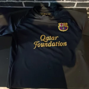 FC Barcelona svart fotbollströja Messi 10 - Säljer en svart FC Barcelona fotbollströja med guldiga detaljer, Messi 10 på ryggen och Qatar Foundation-logga på bröstet. Tröjan har klubbmärket på bröstet och texten 'mes que un club' i nacken. Perfekt för fans!