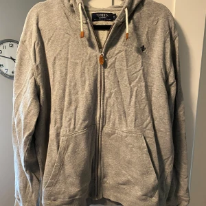 Grå hoodie med dragkedja från Morris - Säljer en grå hoodie från Morris i storlek M. Tröjan har dragkedja, huva med snören och två fickor framtill. Liten broderad logga på bröstet. Perfekt för en avslappnad stil. Hör av dig vid frågor och funderingar!