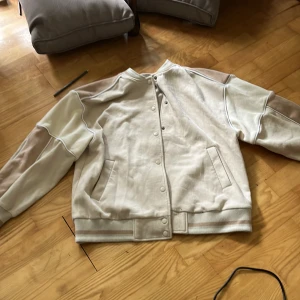 Vit bomberjacka med knappar - Säljer en stilren vit bomberjacka med knäppning framtill och ribbade muddar. Jackan har två fickor på sidorna och en klassisk bomberkrage. Perfekt för dig som gillar en clean och enkel look.