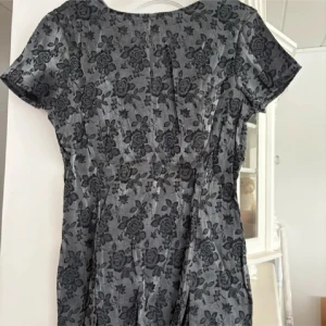 Grå blommig klänning  - Säljer en grå klänning från Lindex med svart blommigt mönster. Klänningen har korta ärmar och en lätt figursydd passform. Perfekt för dig som gillar klassiska och stilrena plagg med en twist.