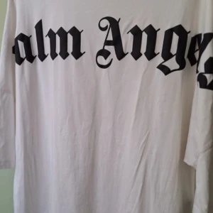Vit t-shirt från Palm Angels - Säljer en vit t-shirt från Palm Angels med stor svart logotyp i gotisk stil både fram och bak samt tryck i nacken. Klassisk passform och rund halsringning. Perfekt för dig som gillar streetwear och vill sticka ut.