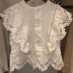Vit spetsblus med öppen rygg - Superfin vit blus med breda spetsdetaljer och vackert broderade kanter. Toppen har korta ärmar med volang och en öppen rygg som knäpps med knapp upptill och nertill. Perfekt för dig som gillar romantisk och bohemisk stil.