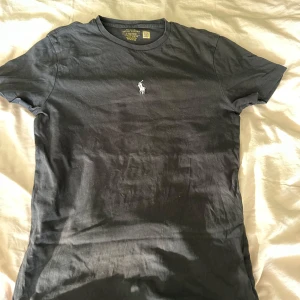 Svart t-shirt från Polo Ralph Lauren - Svart t-shirt från Polo Ralph Lauren med klassisk vit broderad logga på bröstet. Modellen är slim fit med rund hals och korta ärmar. Tillverkad i mjuk bomull som känns skön mot huden. Perfekt för en clean och stilren look. Har små tvätthål, märks inte när man har på den!