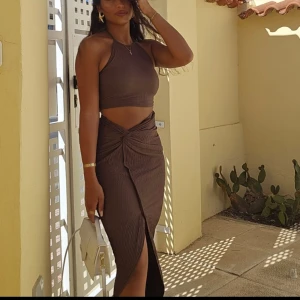 Brun omlott långklänning med slits - Supersnygg brun omlottklänning med ribbad struktur och hög hals. Klänningen är ärmlös och har en cool cut-out i midjan samt en knytning framtill. Lång modell med slits som ger en extra edge. Perfekt för dig som gillar trendiga och unika plagg.