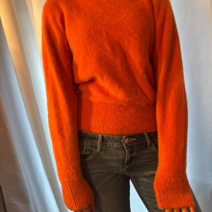 Orange fluffig stickad tröja - Supermjuk och fluffig orange stickad tröja med lång ärm och ribbade muddar. Tröjan har en lite högre hals och sitter snyggt i midjan. Perfekt för dig som vill ha en färgklick i garderoben och gillar en cozy vibe.