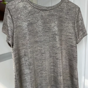 Glittrig silver t-shirt i metallic-look - Snygg t-shirt i silver med glittrig metallic-effekt. T-shirten har korta ärmar och rund halsringning. Materialet är mjukt och skimrande, perfekt för dig som vill sticka ut lite extra. Passar dig som gillar en trendig och cool stil.