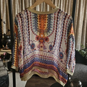 Färgglad blus med guldiga detaljer - Karen by Simonsen - Unik blus med färgstarkt, bohemiskt mönster i blått, orange, rött och gult. Blusen har breda guldiga ribbade kanter vid halsringning och nederkant. Lätt och luftig känsla, perfekt för dig som gillar statement-plagg och snygga detaljer.