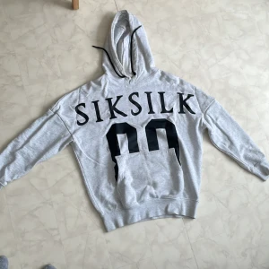 Grå hoodie från SikSilk XS - Snygg ljusgrå hoodie från SikSilk i storlek XS. Stor svart logga och siffror på framsidan, klassisk känguruficka och huva med svarta snören. Den har en liten fläck på insidan av luvan, men går bort enkelt med rätt tvättmedel. För mer frågor kontakta!