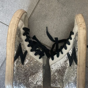 Glittriga sneakers från Le Temps des Cerises - Snygga sneakers med silverglittriga paneler, svarta blixtdetaljer på sidorna och svarta snören. Ovandelen är i vitt syntetmaterial 