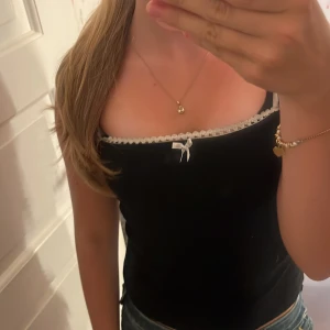 Svart linnetopp med spets från Vero Moda - Supersöt svart linnetopp från Vero Moda med vita smala axelband, spetskant upptill och en liten rosett framtill. Toppen har en figurnära passform och är perfekt för dig som gillar en feminin och trendig look. Tyvärr kommer den inte till användning💞orginal pris 270kr💞