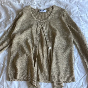 Glittrig guld kofta från Moschino - Säljer en glittrig guld kofta från Moschino Cheap and Chic. Koftan har ribbade muddar, långärmad design och snygga vita knappar framtill. Materialet är stickat med metallictråd som ger en lyxig känsla. Extra detalj med knytband nedtill för en cool look. Står ingen storlek men skulle gissa på xxs