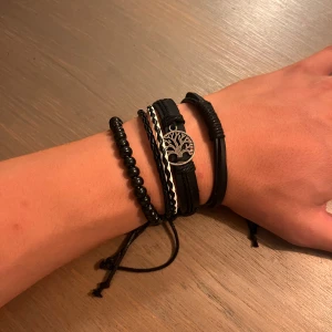 Svart armband med trädberlock - Snyggt svart armband med flera olika band och pärlor, samt en silverfärgad berlock med trädmotiv. Armbandet har både flätade och släta detaljer och justerbar knytning. Perfekt för dig som gillar att mixa stilar och vill ha något unikt på handleden.