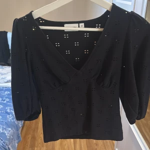 Svart blus med v-ringning från Kappahl - Svart blus från Kappahl i storlek XS med v-ringning och puffiga trekvartsärmar. Toppens tyg har små broderade hålmönster som ger en luftig känsla. Perfekt för dig som gillar stilrena detaljer och en feminin look.