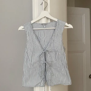 Randig linnetopp från Weekday - Säljer en snygg topp från Weekday i vitt med tunna svarta ränder. Toppen har djup v-ringning och knyt framtill med tre band. 