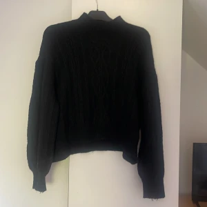 Svart stickad tröja från Samsa Collection - Svart stickad tröja från Samsa Collection med snyggt kabelstickat mönster framtill. Tröjan har långärmade, lite puffiga ärmar och ribbade muddar vid hals, ärmslut och nederkant. Perfekt för dig som gillar en klassisk men chill look.