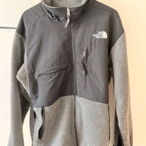 Grå Denali retro fleece från The North Face - Säljer en grå och mörkgrå fleecejacka från The North Face med hög krage och hel dragkedja. Jackan har flera praktiska fickor med dragkedja och är tillverkad i mjuk fleece och syntetmaterial. Perfekt för kyliga dagar och outdoor vibes.