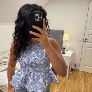 Vit och blå peplum blus med mönster - Superfin blus i vitt och blått med detaljerat mönster och peplum-utsvängd midja. Toppen är ärmlös med breda axelband som korsas i ryggen och har en snygg, figurnära passform. Perfekt för dig som gillar en söt och trendig stil. Är i storlek L men passar mer som S/M.