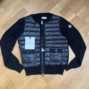 Svart dunväst från Moncler - Snygg svart dunväst från Moncler med quiltad framsida och två stora fickor med knapp. Västen har dragkedja och Moncler-logga på ärmen. Materialmix med glansig polyester framtill och stickade svarta partier på sidor och ärmar. Perfekt för lager-på-lager-stil.