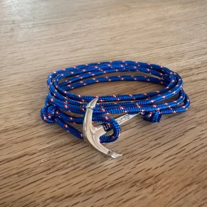 Blå armband med silverfärgad ankare - Snyggt armband med blått, randigt rep och en silverfärgad ankare som lås. Repet har röda och vita detaljer som ger en cool marin känsla. Perfekt accessoar för dig som gillar havet och vill sticka ut med din stil. Ny pris 399