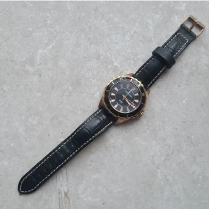 Klocka från Dyrberg/Kern - Stilren klocka från det exklusiva danska märket Dyrberg/Kern med svart urtavla och guldfärgad boett. Klockan har svart läderarmband med vita sömmar och klassisk spännesstängning. Urtavlan har guldiga markeringar och detaljer. Gjord av rostfritt stål och kan även visa datumet. Behövs köpas ett nytt batteri.